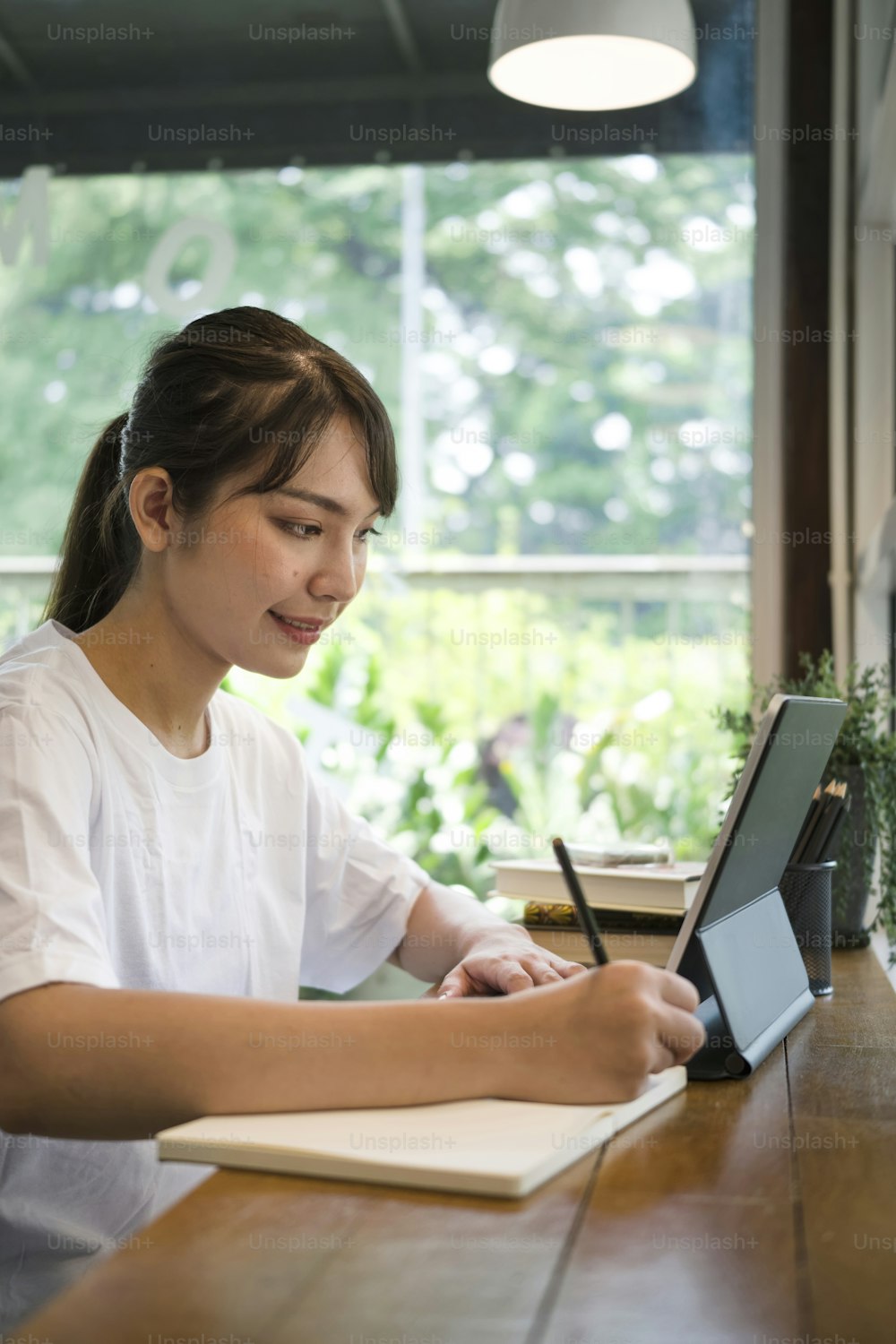 Side view smiling young woman making note on notebook and using computer tablet at coffee shop.