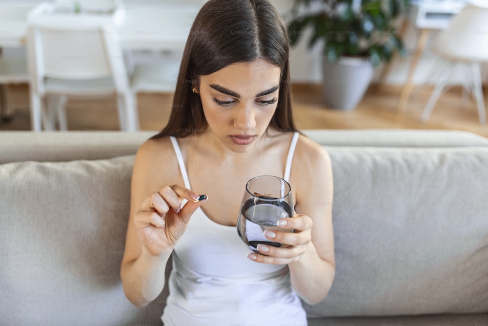 Young woman takes pill with glass of water in hand. Stressed female looking in window and drinking sedated antidepressant meds. Woman feels depressed, taking drugs. Medicines at work