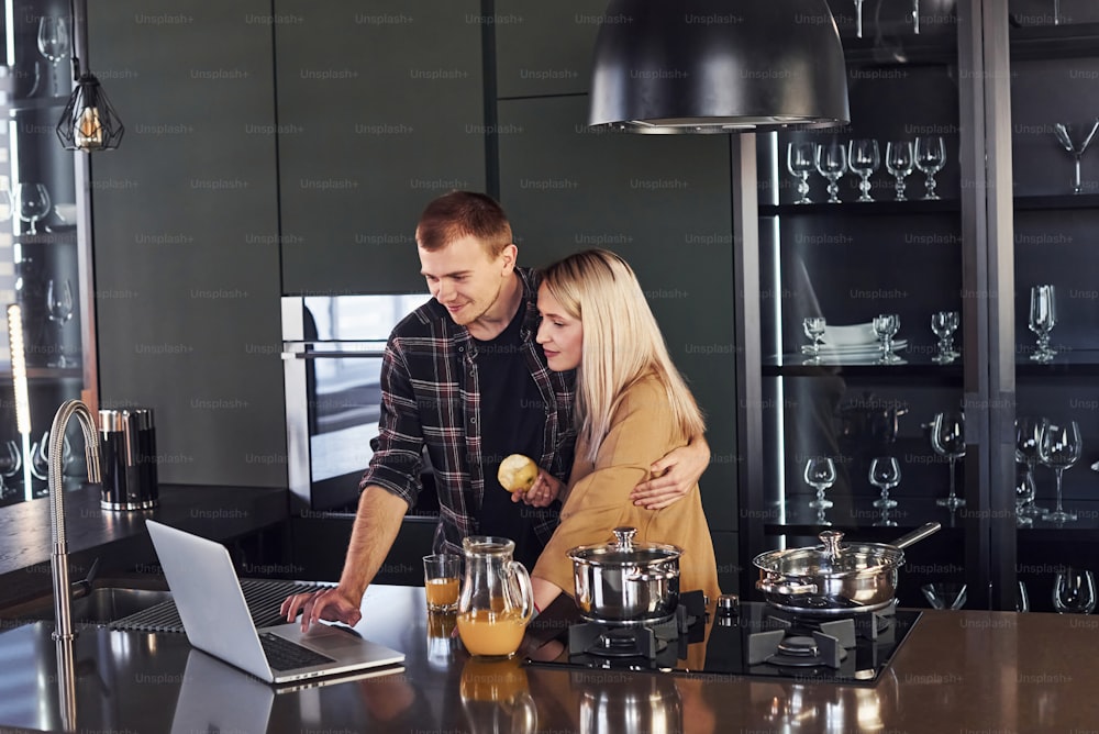 Young couple in casual clothes standing together in kitchen and using laptop.