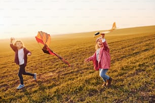Two little girls friends have fun together with kite and toy plane on the field at sunny daytime.