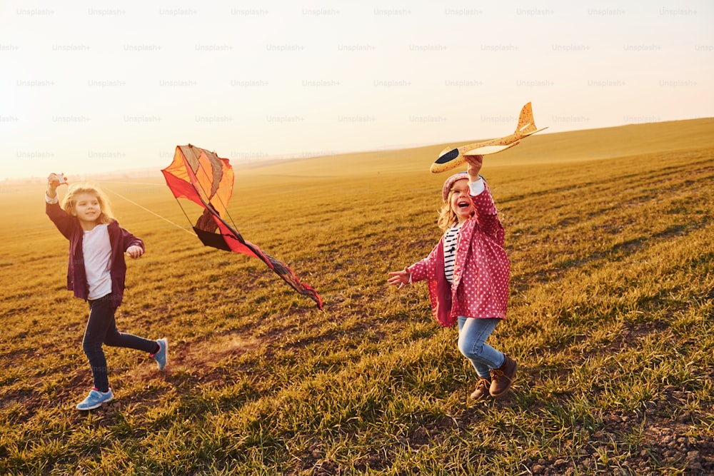 Zwei kleine Freundinnen vergnügen sich zusammen mit Drachen und Spielzeugflugzeug auf dem Feld an sonnigen Tagen.