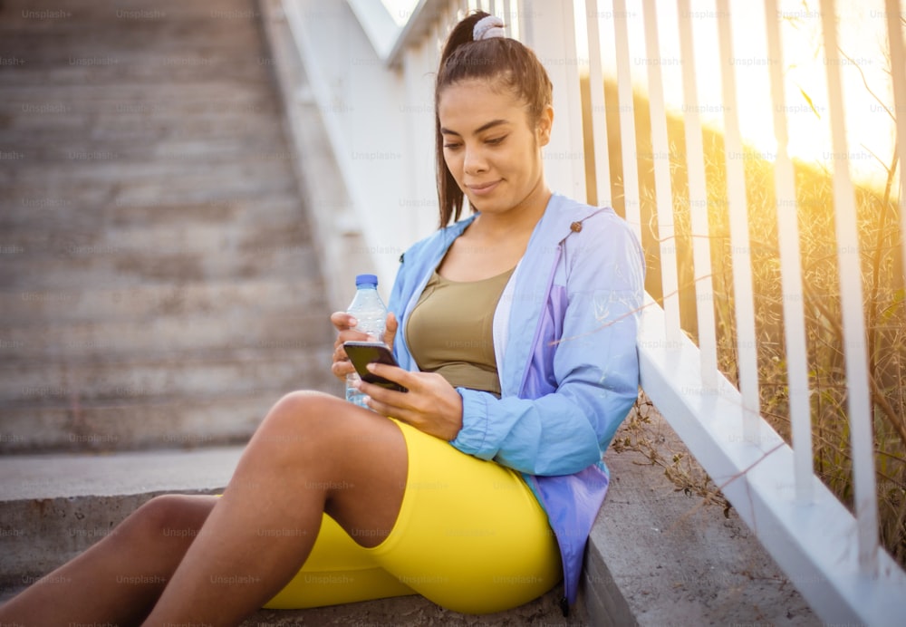 African sports woman sitting on the stairs holding bottle of water and using smart phone.