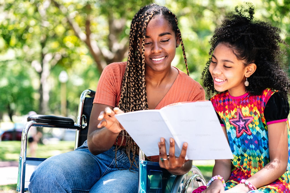An african american woman in a wheelchair enjoying a day at the park with her daughter while reading a book together.