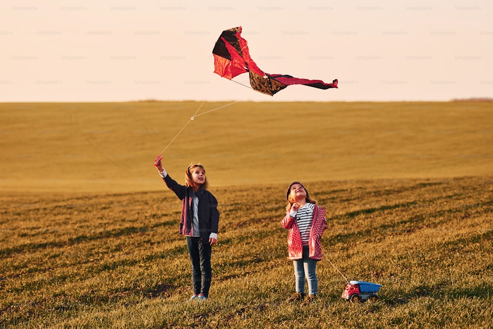 Two little girls friends have fun together with kite and toy car on the field at sunny daytime.