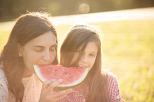 Mother and daughter sitting in nature and eating watermelon.