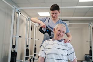 Felice maschio anziano che riceve un massaggio alla spalla da un chirurgo ortopedico, in piedi dietro di lui e usando un massaggiatore a percussione