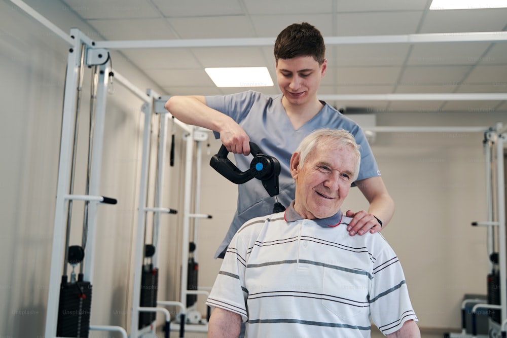 Heureux homme vieillissant se faisant masser les épaules par un chirurgien orthopédiste, se tenant derrière lui et utilisant un masseur à percussion