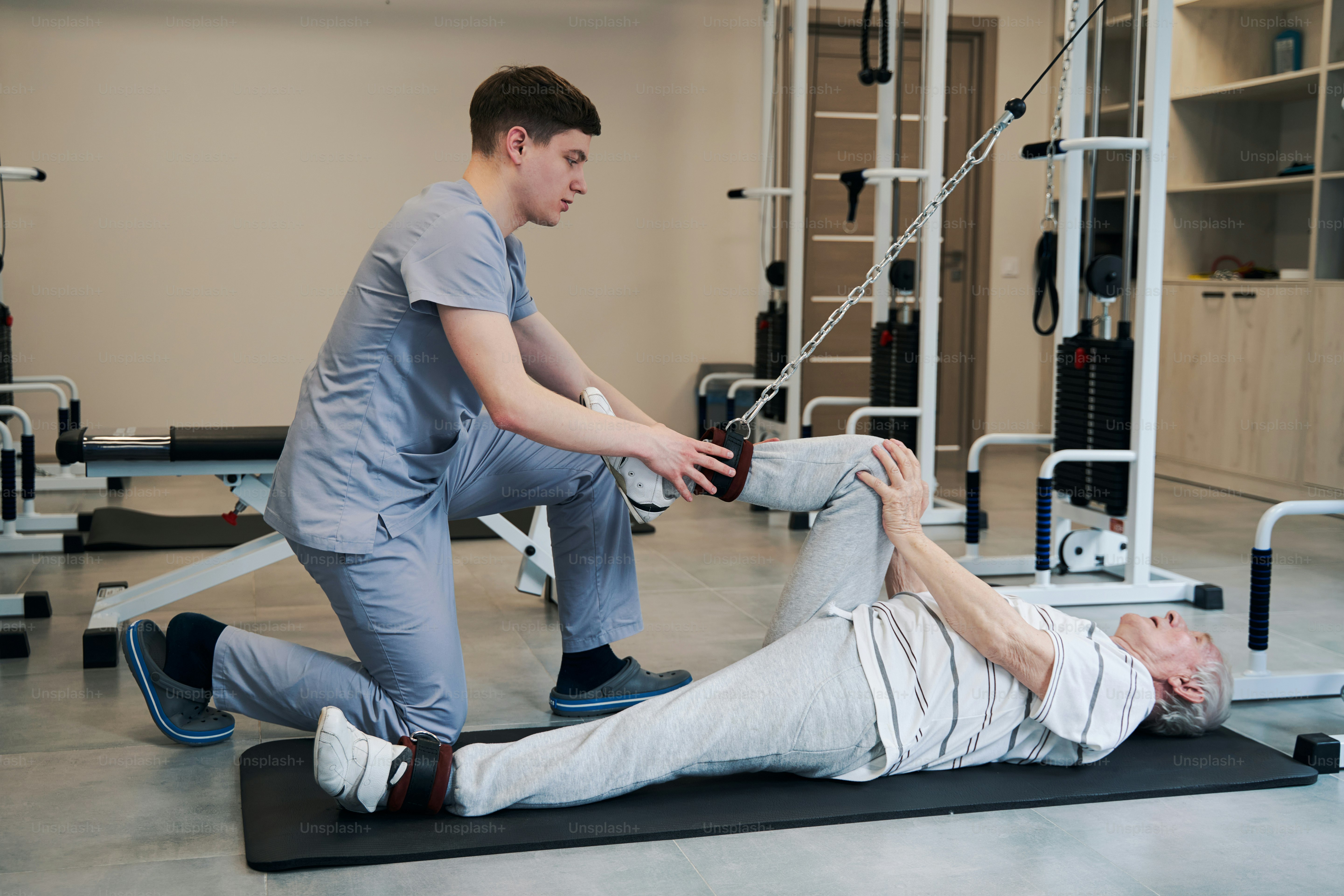 Elderly male working out on fitness resistance training equipment while orthopedic surgeon helping him bend leg