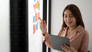 Smiling businesswoman reading sticky notes on glass wall and using digital tablet.