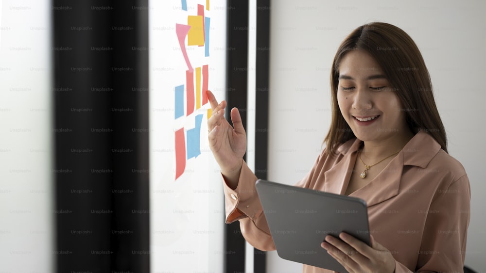 Smiling businesswoman reading sticky notes on glass wall and using digital tablet.