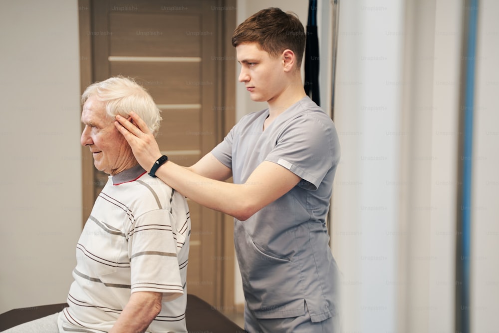 Manual therapist stretching skin of aging man to his ears with hands while standing behind him