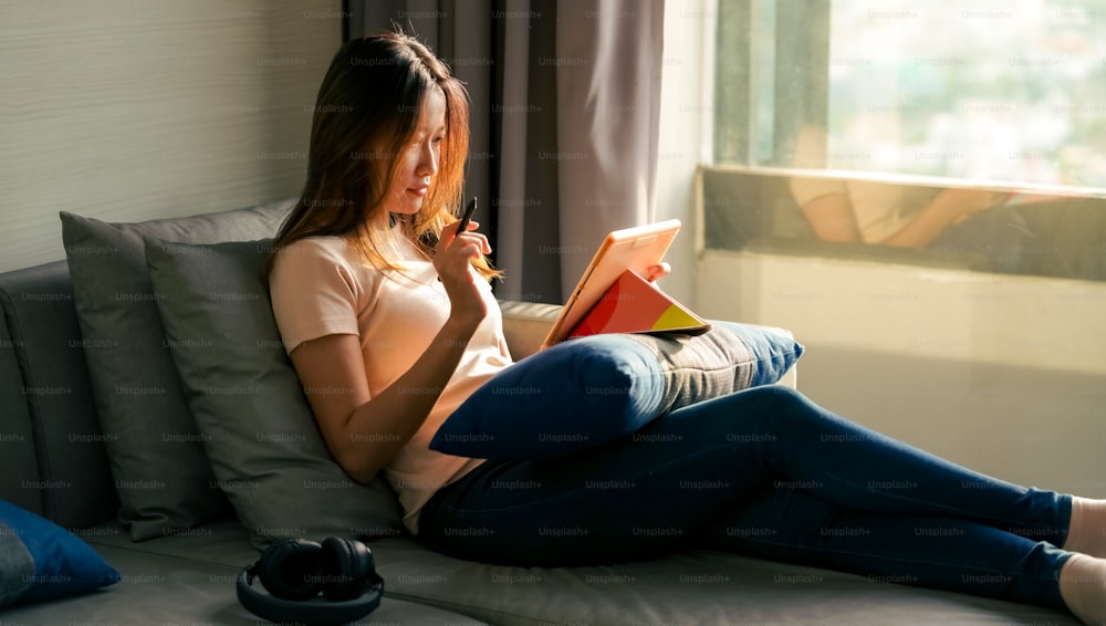 소파에 앉아 아파트에서 온라인 비즈니스를 위해 디지털 태블릿으로 작업하는 아시아 여성. 여성 프리랜서는 집에서 작업 계획이나 온라인 메시지를 위해 태블릿 터치 스크린에 디지털 펜 쓰기를 사용합니다.