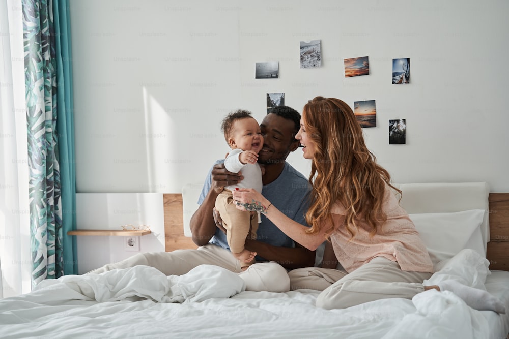 Full length view of the happy friendly family having fun on white bed in the bedroom at the weekend day. Weekend activity, happy family lifestyle concept