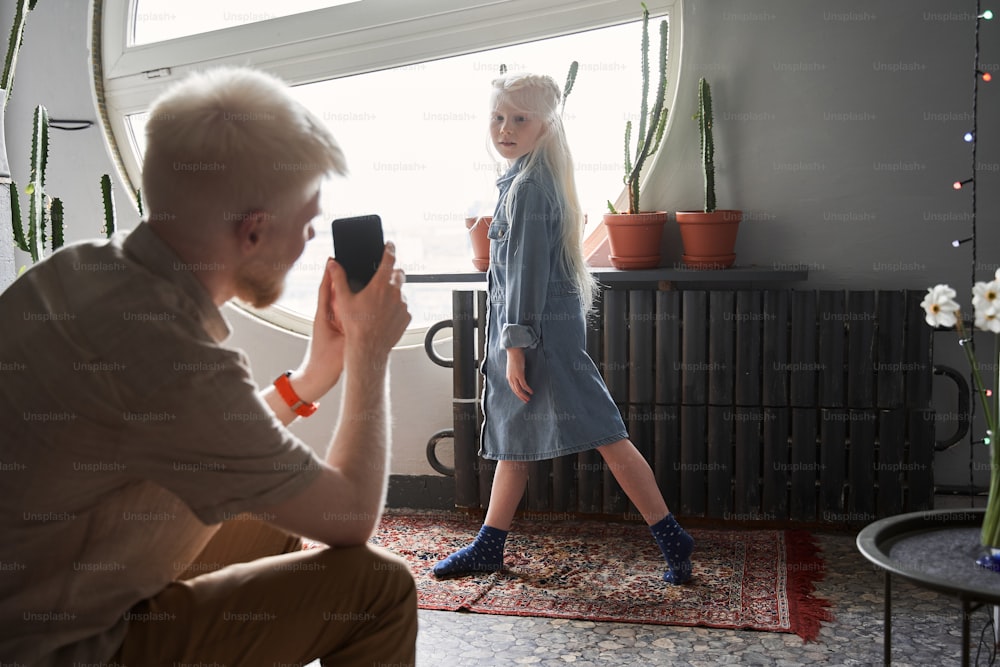 My little princess. Back view of the blonde man shooting at his smartphone his little beautiful daughter while she posing at home. Family relationships concept