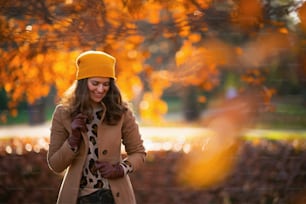 Hello november. happy trendy middle aged woman in brown coat and yellow hat outside in the city park in autumn.