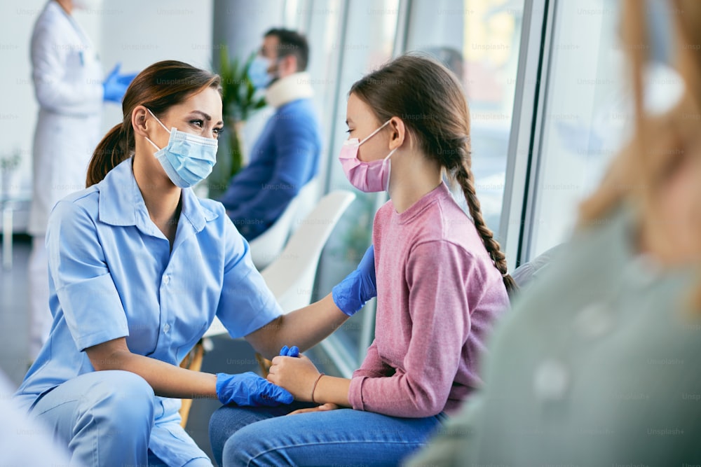 Lächelnde Krankenschwester und kleines Mädchen mit schützenden Gesichtsmasken, während sie im Flur des Krankenhauses kommunizieren.