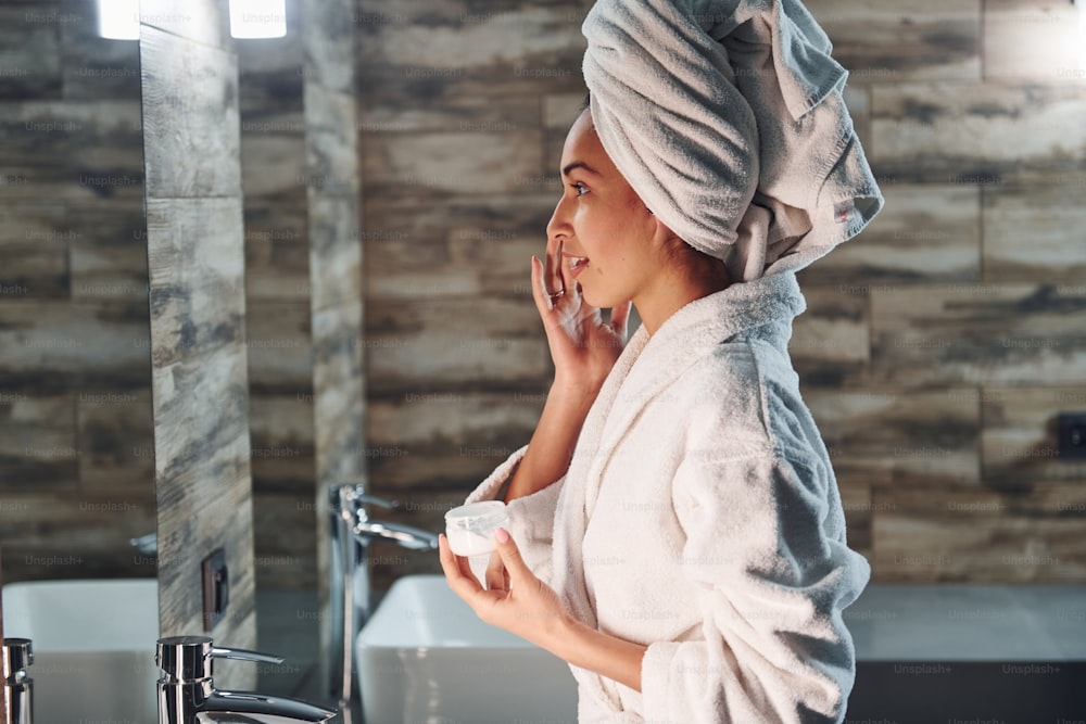 Beautiful young woman in white towel standing in bathroom and taking care of her face.