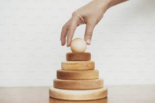 Eco friendly plastic free modern toys for toddler. Hand holding natural wooden pyramid ring on table on white wall background. Montessori education and play. Stylish simple wooden toy for child