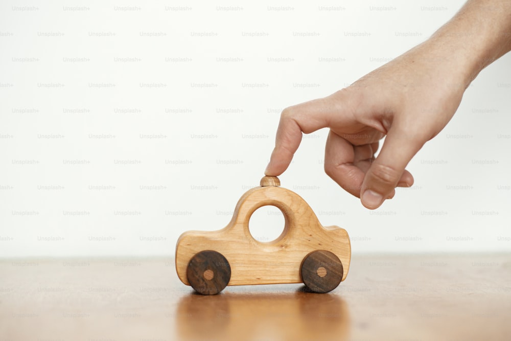 Road trip and travel concept. Hand playing with natural wooden car toy on table on white wall background. Space for text. Eco friendly plastic free toy for toddler. Stylish simple toy for child