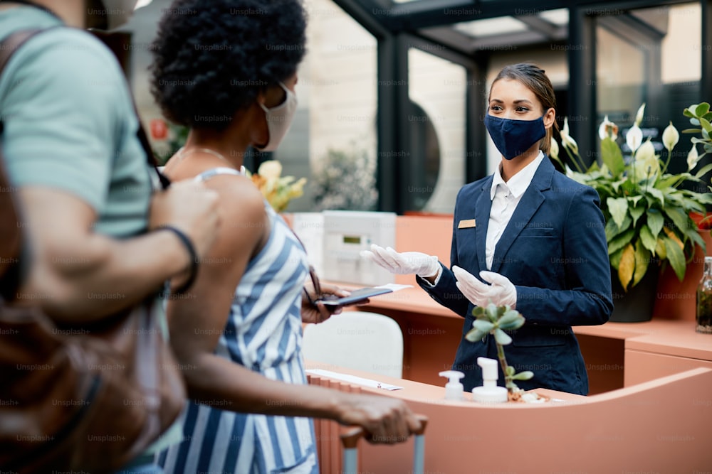 Felice receptionist dell'hotel che indossa una maschera protettiva per il viso mentre comunica con gli ospiti durante il check-in.