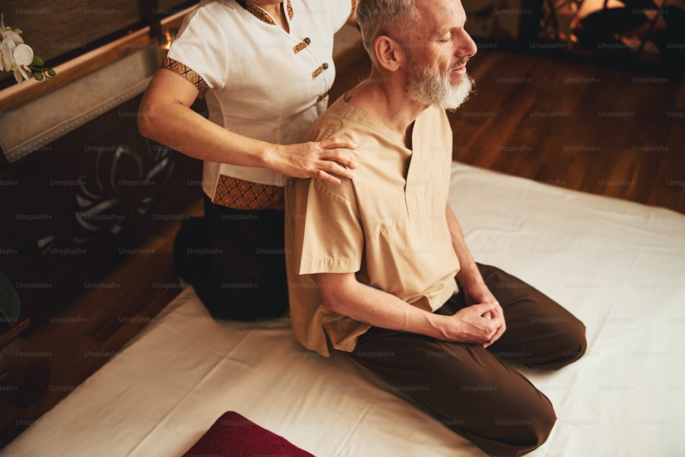 전문 여성이 수동으로 어깨를 치료하는 동안 무릎을 꿇고 앉아 있는 편안한 회색 남성의 상위 뷰