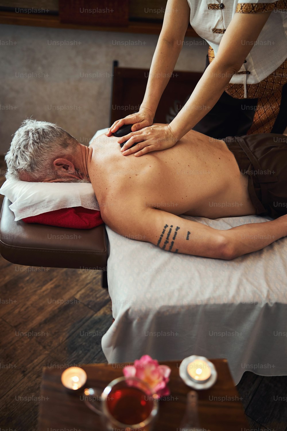 Top view of grizzled tattoed male lying on couch while Asian woman is treating his back with stones