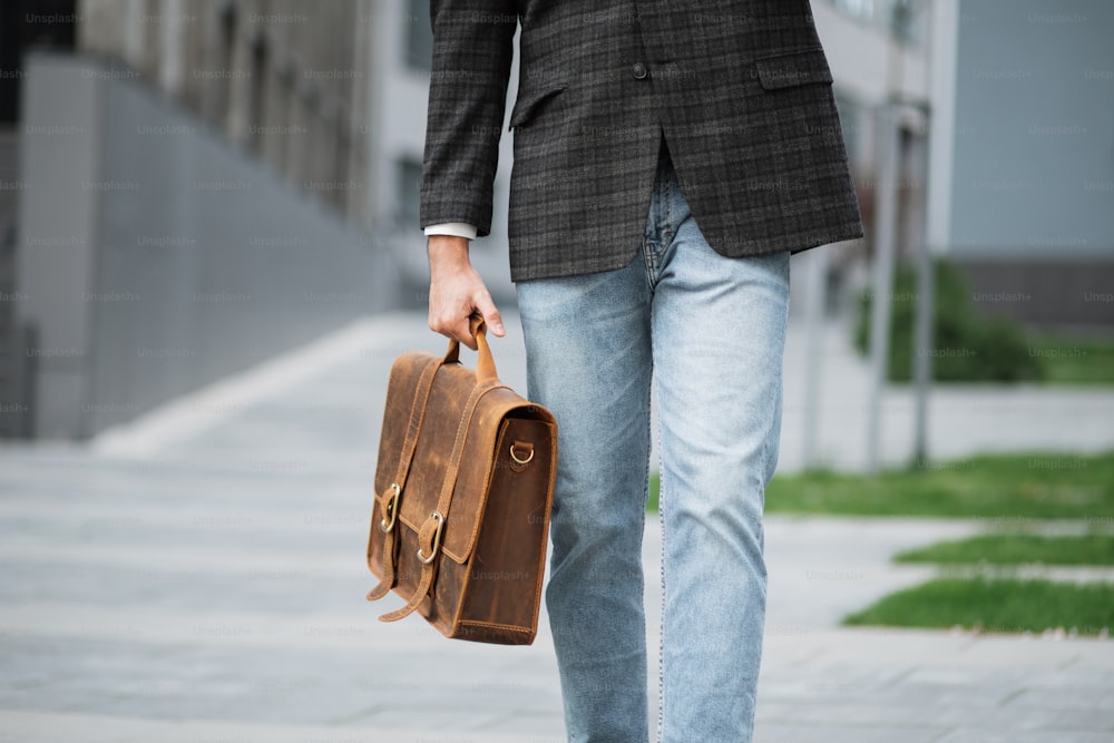Close up of young man dressed in blue jeans and formal jacket carrying brown leather suitcase while walking near office building. Concept of successful people, business and career.