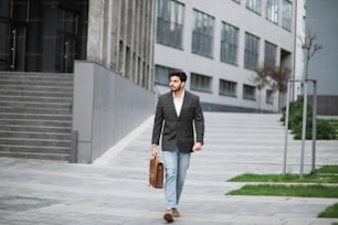 Handsome muslim businessman in stylish formal wear walking outdoors with brown leather suitcase in hands. Bearded young guy on way to his work.