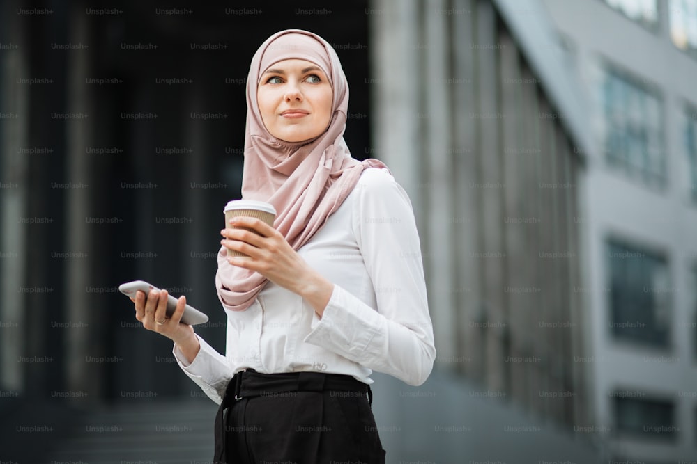 オフィスセンターの近くに立ち、モダンなスマートフォンとコーヒーカップを持っている魅力的なアラビア人女性。フォーマルな服を着てヒジャブをかぶった自信に満ちた女性が、路上でポーズをとっている間、脇を見ています。