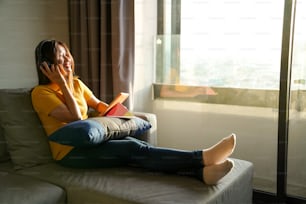 Asian woman sitting on sofa listening to the music from headphones while online working on digital tablet for business project at apartment. Female freelance work job plan or send online message at home.