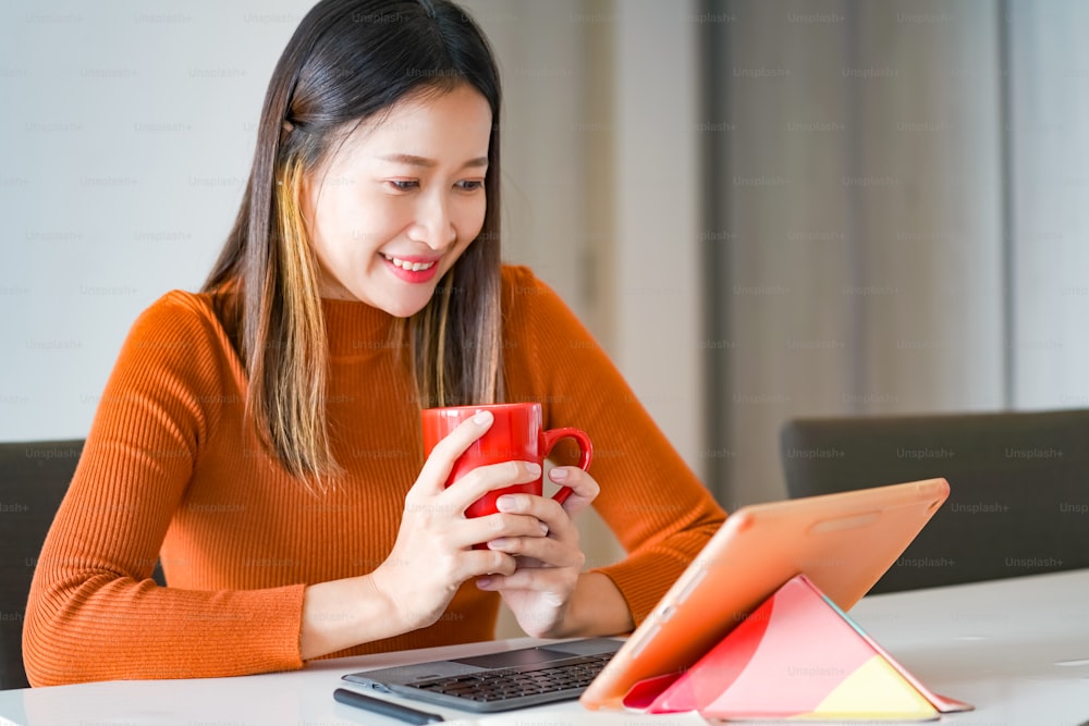 Asian woman using digital tablet with internet for online small business work on table in apartment. Female freelance typing and scratching on tablet touch screen for working business job or online shopping at home.