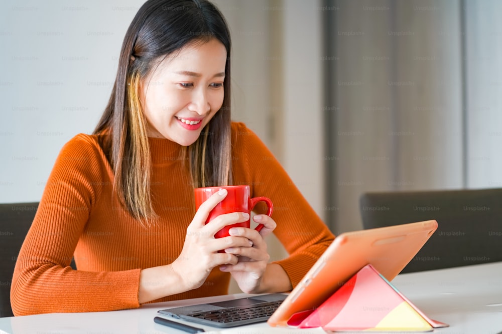 아시아 여성은 아파트의 테이블에서 온라인 중소기업 작업을 위해 인터넷과 디지털 태블릿을 사용합니다. 여성 프리랜서는 비즈니스 업무나 집에서 온라인 쇼핑을 하기 위해 태블릿 터치 스크린에 타이핑하고 긁습니다.