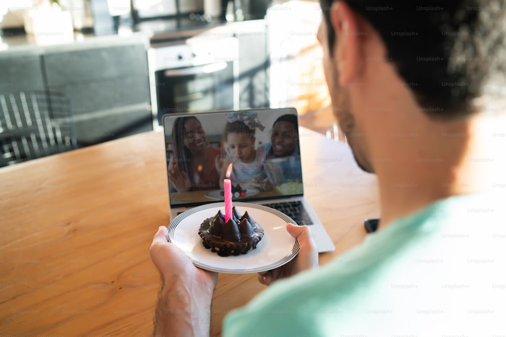Portrait of young man celebrating birthday on a video call from home with laptop and a cake. New normal lifestyle concept.
