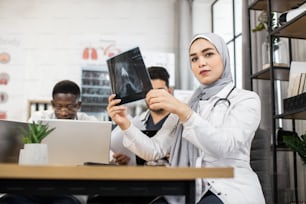 Team of international competent doctors discussing results of x ray scan. Multiethnic medical workers sitting at conference room and using laptops with tablets. Muslim lady in hijab looks at the x-ray
