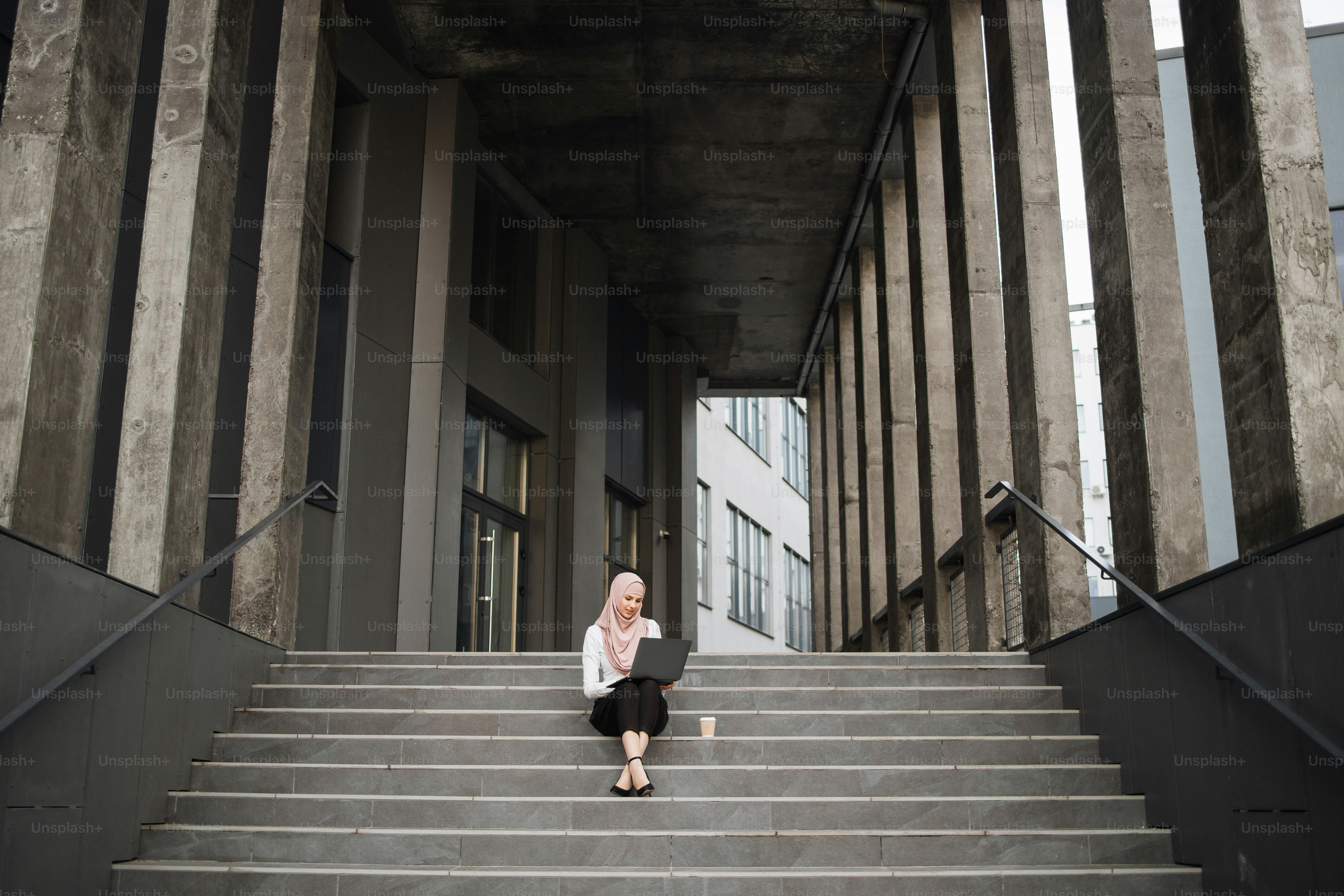 Junge muslimische Frau, die auf einer Treppe in der Nähe des Business Centers sitzt und an einem drahtlosen Laptop arbeitet. Hübsche Frau in eleganter Kleidung und Hijab. Freiberufliches Konzept.