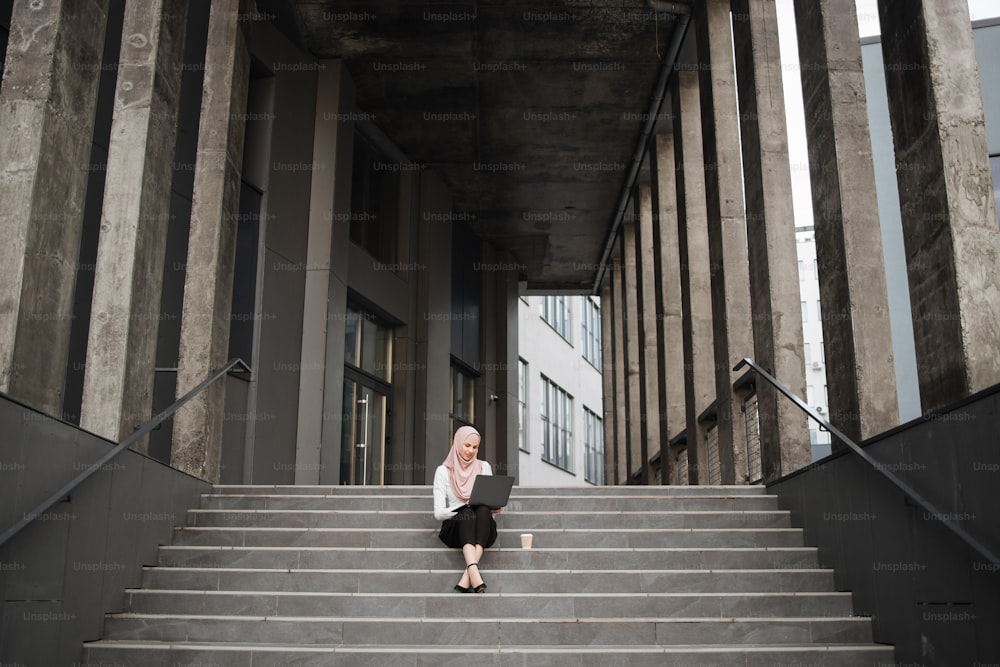 Young muslim woman sitting on stairs near business center and working on wireless laptop. Pretty woman wearing elegant clothes and hijab. Freelance concept.