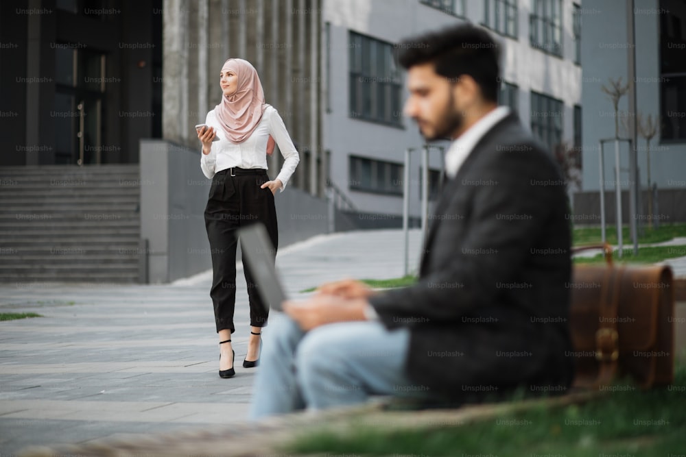 Unscharfer Vordergrund des muslimischen Mannes in stilvoller Kleidung, der auf der Bank sitzt und am Laptop arbeitet. Hübsche Frau im Hijab läuft auf dem Hintergrund mit Smartphone an den Händen. Stadtgebiet.