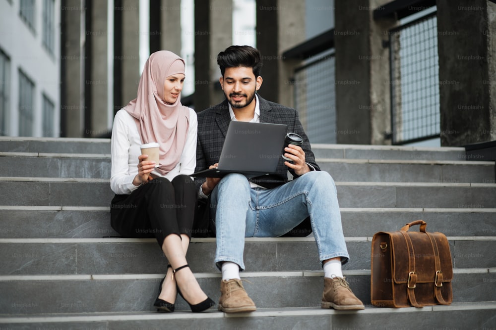 Stilvoller muslimischer Mann und charmante Frau im Hijab, die zusammen auf der Treppe sitzen, lächeln und reden. Zwei Freiberufler, die einen Laptop für die Arbeit im Freien verwenden.