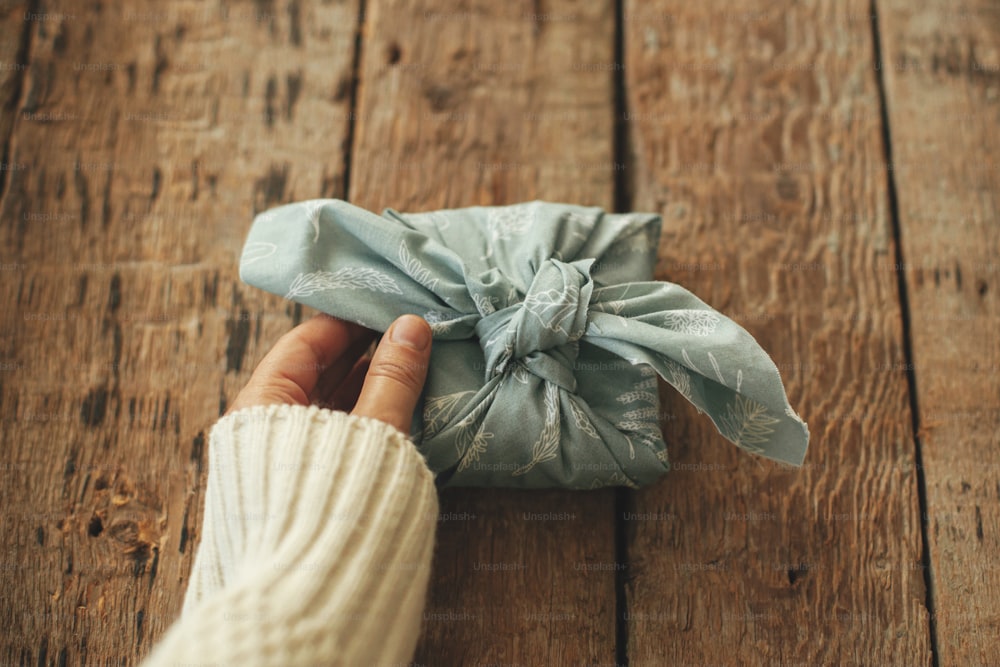 Furoshiki umweltfreundliches Geschenk. Hand hält stilvolles Geschenk in Stoff verpackt auf rustikalem Holztisch. Frohe Feiertage. Zero Waste Winterurlaub, Geburtstag oder Muttertag