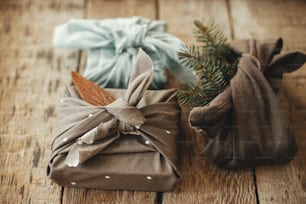 Zero waste stylish christmas gifts. Xmas presents wrapped in fabric with fir branch and leaf on rustic wooden table. Eco friendly winter holidays. Furoshiki present. Happy holidays