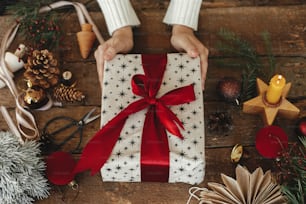 Hands holding stylish christmas gift wrapped in paper on rustic wooden table with festive holiday decorations, top view. Xmas present and candle, scissors, pine cone, fir branches, star