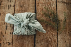 Zero waste winter holidays and gift. Furoshiki eco friendly present. Stylish christmas gift wrapped in fabric and fir branch on rustic wooden table flat lay. Happy holidays and Merry Christmas