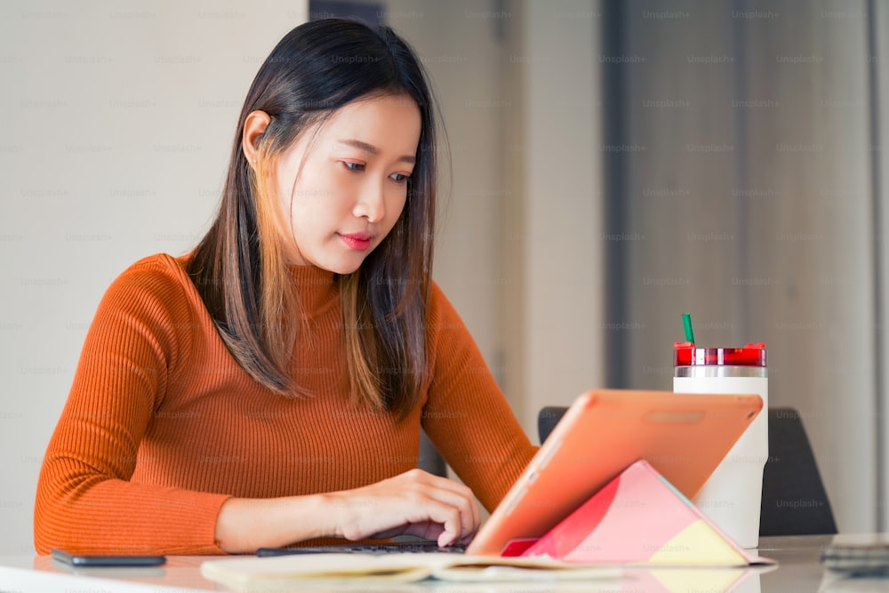 아시아 여성은 아파트의 테이블에서 온라인 중소기업 작업을 위해 인터넷과 디지털 태블릿을 사용합니다. 여성 프리랜서는 비즈니스 업무나 집에서 온라인 쇼핑을 하기 위해 태블릿 터치 스크린에 타이핑하고 긁습니다.