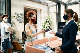 Female tourist with protective face mask giving passport to receptionist while checking in a hotel with her boyfriend.