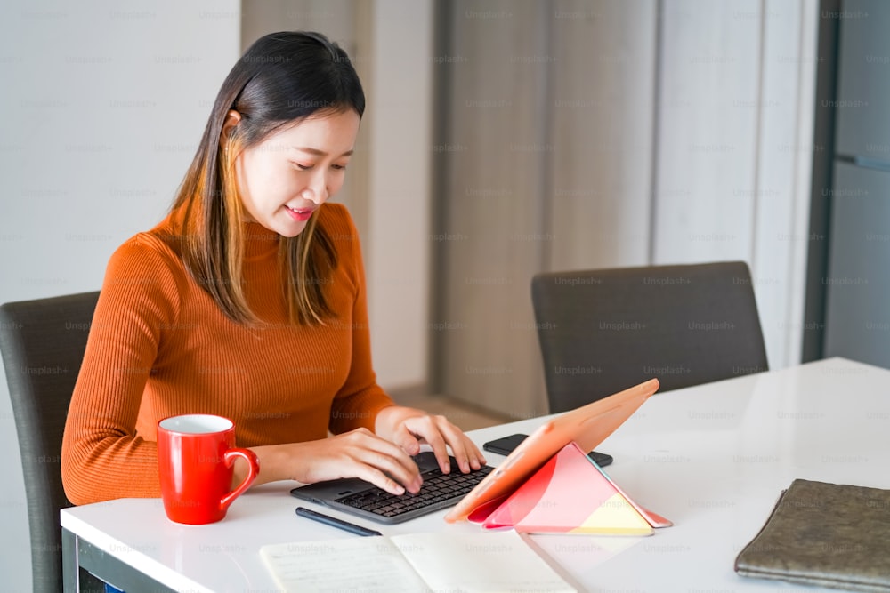 Asian woman using digital tablet with internet for online small business work on table in apartment. Female freelance typing and scratching on tablet touch screen for working business job or online shopping at home.
