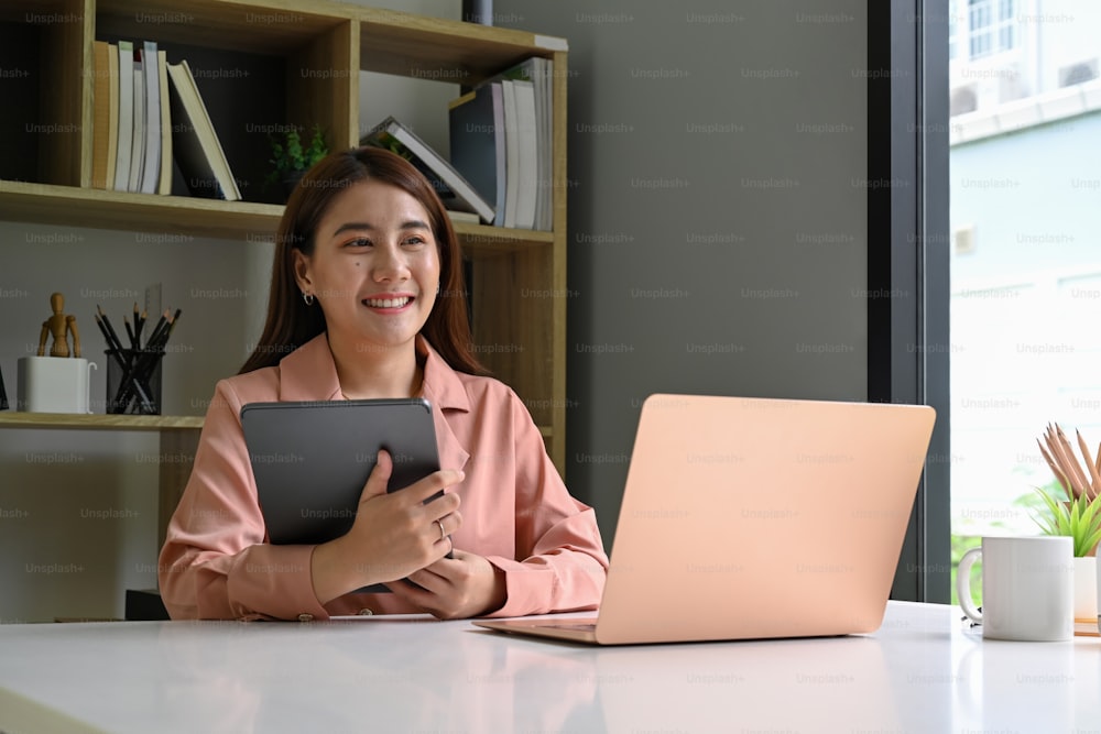 Smiling young woman holding digital tablet and sitting at office desk.