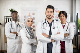 白い白衣を着た多民族の医師のチームが、医療センターで一緒に立っている間、手を交差させています。人、医学、チームワークのコンセプト。