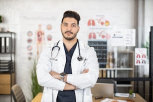 Portrait of confident Arabian male doctor keeping in white lab coat posing at hospital room with his arns crossed folded. Concept of people, health care and treatment.