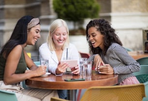 Tres mujeres sentadas en un café en la calle y hablando. Usando un teléfono inteligente.