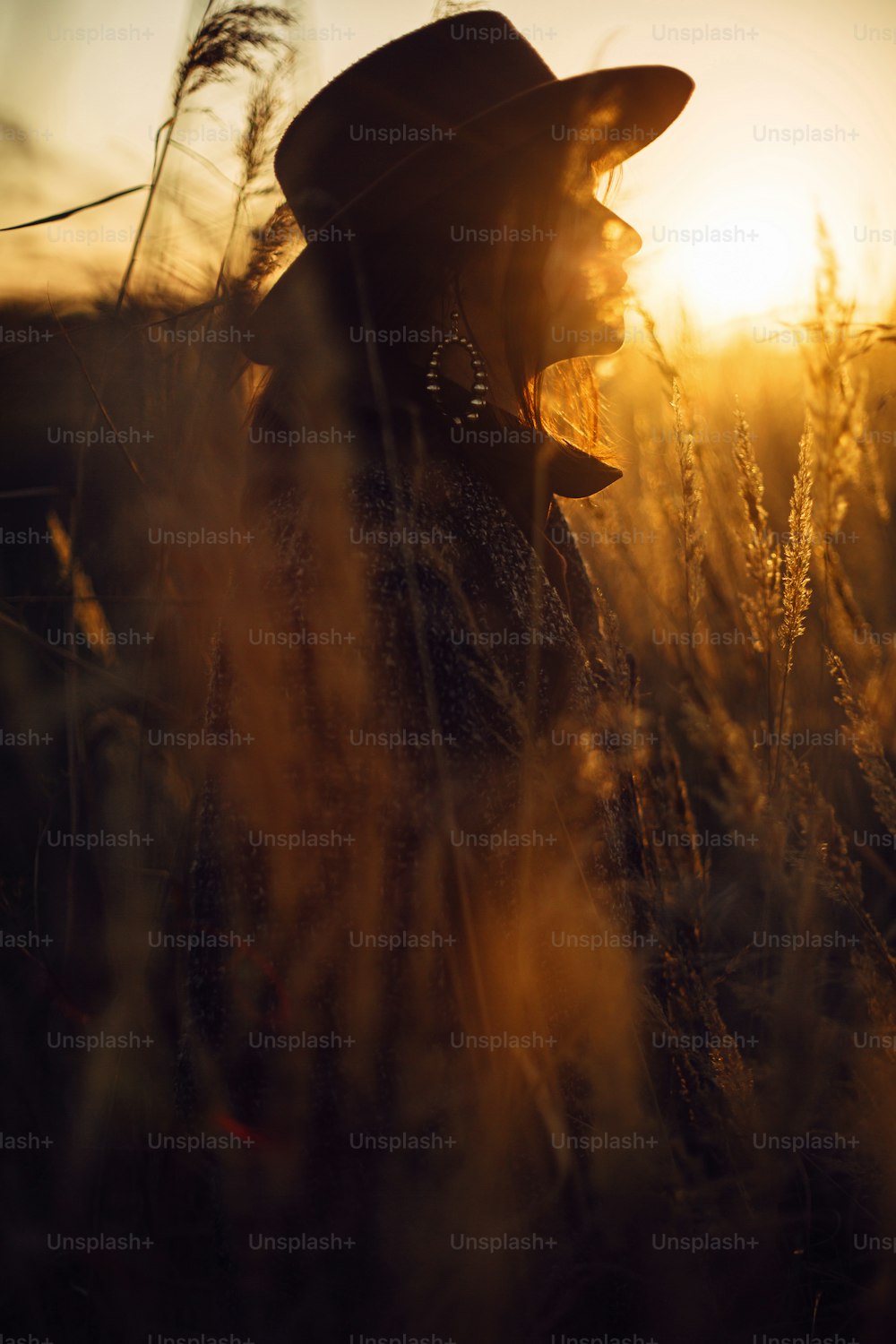 秋の野原の暖かい夕焼けの光の中で草むらに立つ帽子をかぶった美しいスタイリッシュな女性のシルエット。静寂に包まれたひととき。田舎で夜を楽しむファッショナブルな若い女性
